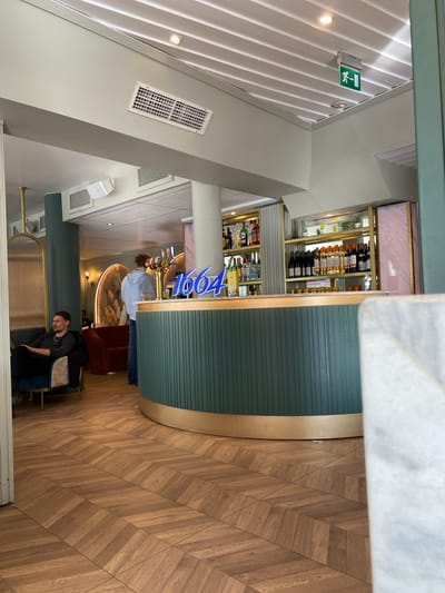 The image depicts the interior of a bar/restaurant in Uppsala, Sweden. The dominant feature is a curved bar with a blue-green, horizontally-ridged facade and a golden trim. Shelves behind the bar are stocked with bottles of alcohol. A blue neon sign that reads "1664" is seen above the bar. The floor is made of light brown wood planks arranged in a herringbone pattern. In the foreground, a man wearing a grey shirt and dark pants is seated. Two people are seen standing at the bar. The ceiling is white with linear indentations and several air vents. An exit sign is visible on the left.