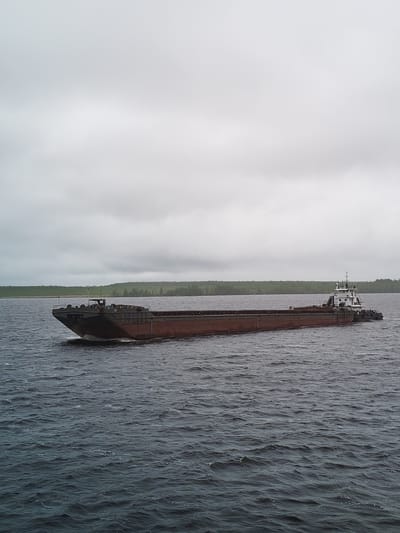 A long, low barge, brown and black, is seen moving on a body of water under an overcast sky. The barge is being pushed or pulled by a smaller tugboat at its rear. In the background, a line of trees is visible along the shore. The location is Gorushka, Russia. The body of water has small waves.
