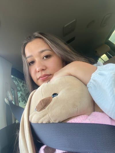 A woman is in the passenger seat of a vehicle, holding a stuffed animal. She is wearing a light-colored top with ruffled sleeves. The stuffed animal is brown and appears to be a bunny. A seatbelt is visible across both the woman and the stuffed animal. The background shows the interior of the car, including the ceiling and a portion of the driver's seat. Outside the car window, trees are visible. The lighting suggests it is daytime. The location of Staromusino, Russia is not visible or inferable from the image.