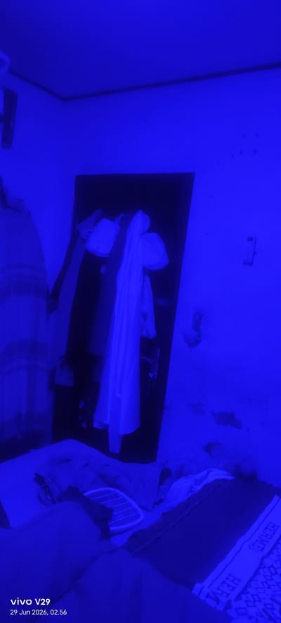 The image depicts an interior scene illuminated by a strong blue light, creating a monochromatic blue environment. The focus is on a doorway with several white garments hanging on or in front of it, one of which is a long, flowing dress. To the left of the doorway, a striped garment is visible. In the foreground, a bed is covered with bedding, and a pair of striped slippers or sandals are placed on top of the covers. The wall to the right of the doorway shows some texture and discoloration. Text at the bottom left corner of the image reads "vivo V29 29 Jun 2026, 02:56", indicating the device and timestamp of the photo, suggesting it was taken in a residential setting. The location specified is Pulodolang, Indonesia.