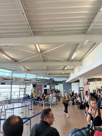 An indoor scene, likely a busy airport or transit terminal in Tillé, France, captured in the afternoon at 14:43. The image shows a wide waiting area with numerous people.

In the foreground, a man's bald head and shoulder are visible from behind, facing away from the camera. Slightly to his left, another man with glasses and dark hair is seen in profile, looking towards the queue area. The mid-ground is dominated by several rows of stanchions with blue retractable belts, forming a queue line where a few people are standing. Beyond the queue, many individuals are seated in metal waiting chairs.

A woman with brown hair, wearing a black top and a green wristband, is centrally located, looking directly at the camera while talking on a mobile phone. Further back, another woman in a long black dress and olive green jacket, carrying a backpack, stands among the crowd. A "Reebok" bag is visible near some seated passengers.

The building features a high, corrugated metal ceiling with exposed structural beams and fluorescent lighting. Large windows run along one side of the terminal, providing natural light, below which air conditioning units are mounted. Green signage reading "Café kiosque" and "café bosque" is partially visible in the background, indicating refreshment services. A digital clock clearly displays "14:43," and a sign with a red arrow points to "PORTES GATES D-E," confirming an airport setting. The floor is tiled in a light beige color. The overall atmosphere appears to be one of people waiting patiently in a modern, functional public space.