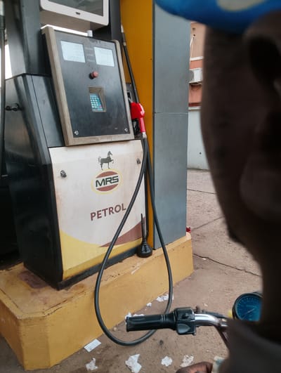 This media file captures a close-up view of a fuel pump at a gas station, likely in Isheri, Nigeria. The primary object is a petrol pump with a visible display screen showing "0.00 NAIRA," "0.00 LITRE," and "PRICE 1205.00." Below this, a keypad and a red button are visible. The pump's exterior is white and yellow, with a logo featuring a horse and the text "MRS PETROL" prominently displayed. A black fuel hose with a red nozzle is attached to the pump.
The scene is viewed from the perspective of someone on a motorcycle, as evidenced by the handlebars and a portion of the motorcycle's dashboard, which shows a blue-faced speedometer with a needle pointing around 20-30. A person's hand, holding a banknote, is also visible, suggesting a transaction or an impending one. The background shows a light brown, paved ground with scattered white paper debris and cracks. A building with a light-colored wall and a white air conditioning unit is partially visible in the distance.
The lighting suggests it is daytime, with no specific weather conditions discernible. The overall impression is a typical, everyday scene at a Nigerian gas station, focusing on the fuel dispensing equipment and the immediate experience of a customer. The text "MRS PETROL" and the numbers on the display are the only clearly legible text in the image.