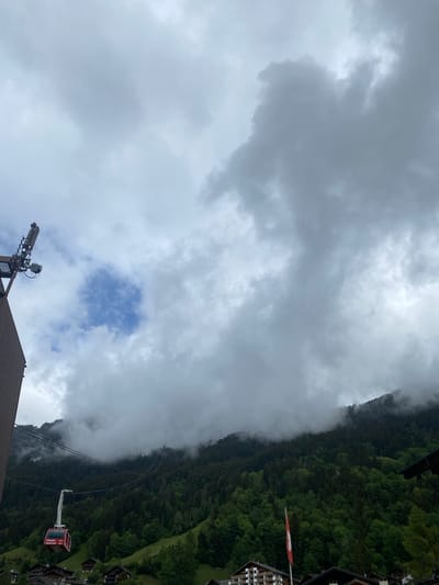 The image displays a mountainous landscape under a predominantly overcast sky, with a small patch of blue visible through the clouds. A dense, dark green forest covers the steep mountain slopes, which are partially enveloped in low-hanging clouds or fog, giving the scene a misty atmosphere.
In the foreground, the roof of a building is visible, featuring a prominent metal mast equipped with various antennas and communication dishes. To the right, another building, likely an alpine chalet, has a red and white Swiss flag flying from it. A red gondola cabin, part of a cable car system, is seen suspended on wires, moving up or down the forested mountain. The gondola cabin clearly displays the text "Champéry" and "Portes du Soleil", identifying the location as a part of this well-known ski region in Switzerland. Several other wooden alpine buildings are nestled among the trees further up the slope.
The scene captures a daytime moment with subdued lighting due to the heavy cloud cover, suggesting cool and potentially damp weather characteristic of mountain environments. The overall impression is one of a functional mountain resort amidst natural beauty and varying weather conditions.