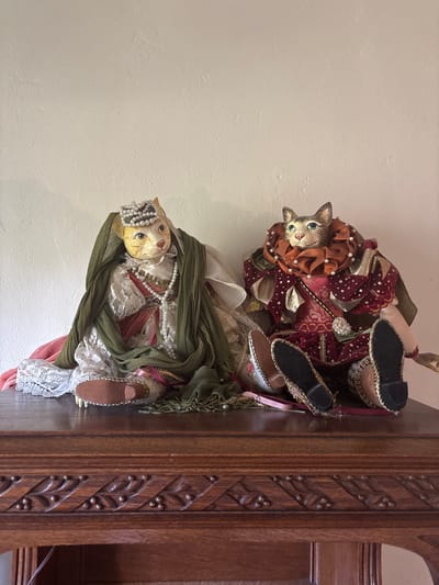 The scene depicts an indoor display in Monterey, United States of America. Two elaborately dressed feline figurines are seated side-by-side on a dark, carved wooden surface, possibly a chest or table, against a plain, light-colored wall.

The figurine on the left features a light-colored cat face with green eyes, wearing a pearl-encrusted crown. It is dressed in a multi-layered garment, including a white lace gown with a red or pink underdress, adorned with a long strand of pearls. An olive-green sheer fabric is draped over its shoulders and lap. Its feet wear soft, pointed shoes.

The figurine on the right also has a light-colored cat face with prominent green eyes, gazing slightly upward. It wears a large, ruffled, burnt-orange collar embellished with pearls. Its attire is a rich red garment with gold patterns, white polka dots, and gold accents, suggesting a historical or theatrical style. Its feet are extended forward, displaying the soles of its pointed, embroidered shoes, one appearing black-soled and the other tan/gold-soled.

The wooden surface supporting the figurines features a carved decorative frieze along its visible edge, comprising repeating leaf-like motifs and small circular indentations. The wall behind is unadorned and has a subtle textured finish. The lighting is even, suggesting an interior space.