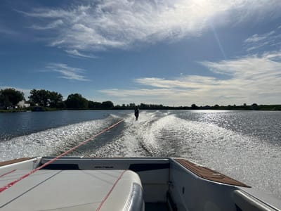 The image captures an active water sports scene on a body of water in Coruche, Portugal, under a bright, partly cloudy sky. The perspective is from the aft section of a white and blue Nautique brand speed boat, which is in motion, producing a substantial twin wake. A single individual is water-skiing approximately 20-30 meters behind the boat, positioned slightly to the right of the centerline of the wake. The individual is wearing dark clothing and holding a red tow rope that extends from the stern of the boat. The water surface reflects bright sunlight, particularly on the right side of the frame. The left shoreline features green trees and a white building with a blue roof. The right shoreline appears as a distant, low-lying landmass with sparse vegetation. The sun is visible as a bright glare in the upper right sky quadrant.