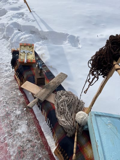 This image, captured in Plast, Russia, depicts an outdoor winter scene featuring a display of traditional items arranged on a makeshift snow-covered surface. The primary subject is a low wooden bench-like structure, draped with a dark plaid blanket featuring red, orange, and yellow patterns with fringed edges. This structure is positioned in deep, undisturbed snow under bright daylight conditions, casting subtle shadows.
On the blanket, from left to right, are several objects:
1. An ornate, reddish-orange wooden box with painted floral designs, containing a quantity of finely shredded or grated light-yellow material, possibly butter or cheese. A decorative silver utensil with a stylized handle rests within this material.
2. An antique cast-iron clothes iron, dark-colored with a black handle.
3. Two wooden paddles or boards; the left one is lighter-toned and appears smoother, while the right one is darker, more weathered, and possibly a *valek* (laundry paddle).
4. Bundles of natural-colored wool or yarn. A large, shaggy bundle of mottled grey and white fibers rests on the blanket, adjacent to a neatly wound ball of light grey yarn.
5. A slender, carved wooden spinning tool or spindle with several dark bands, positioned next to the yarn ball.
6. To the far right, a weathered, light blue wooden object, possibly a frame or part of a larger structure, is partially visible.
7. Above the blue object, a loose mass of dark brown, shaggy fibers, resembling unspun wool or animal hair, hangs from an upright, natural wooden stick or branch.
The ground to the left of the bench reveals a section of a faded red and black patterned rug or textile, partially obscured by a light dusting of snow and ice. The background consists entirely of expansive, undisturbed snowdrifts under a clear sky. No individuals are present, and no active interactions are depicted.
