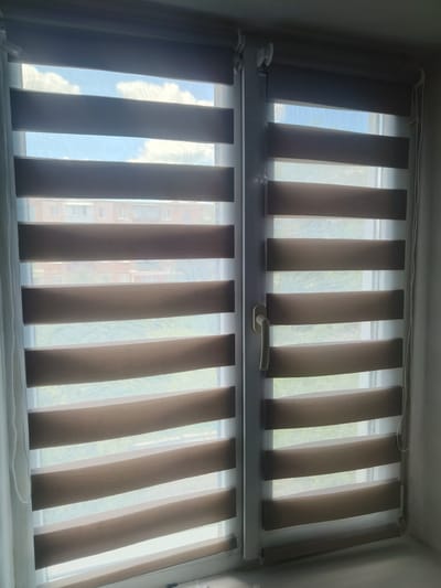 This is a close-up, eye-level shot of a window with zebra blinds, located in Kalynivka, Ukraine. The blinds are partially open, allowing light to filter through. The blinds have alternating horizontal stripes of opaque brown fabric and sheer white fabric. Through the sheer stripes, a blue sky with white clouds is visible, along with the upper floors of a building. Through the opaque stripes, the details of the building are obscured, but it appears to be a multi-story residential building with multiple windows. The overall scene suggests an interior view from a room looking out onto a typical urban landscape in Ukraine.