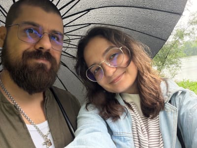 A man and a woman are captured in a selfie under a black umbrella on a rainy day. The man, on the left, has a beard and wears glasses and a green jacket. A silver chain with a cross pendant is visible around his neck. The woman, on the right, also wears glasses and a light blue denim jacket over a striped shirt. They are positioned in the foreground, with the umbrella and raindrops prominently displayed. The background suggests an outdoor setting, possibly near a body of water, with trees and vegetation visible. The image is taken from a slightly elevated angle, looking down on the subjects. The weather is overcast and wet, as indicated by the rain on the umbrella and the overall muted lighting. The location is identified as Nikulskoye, Russia.