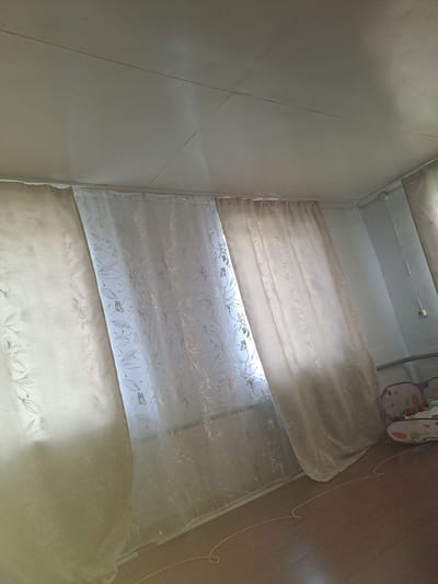 The image shows a room with curtains covering a window. The curtains are of two types: a sheer white curtain with a floral and butterfly pattern, and a thicker, opaque beige curtain. The ceiling appears to be made of drywall and has visible seams and cracks. The floor is made of wood. In the background, a small part of a child's seat with colorful patterns is visible. There are no people visible in the image. The location is a room in the city of Стрельцы, Russia.
