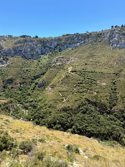 The image shows a steep, rocky hillside in the Italian countryside, possibly near Noto, Sicily. A winding, narrow dirt path zigzags its way up the incline, disappearing into the dense green vegetation that covers much of the slope. The upper parts of the hill are characterized by sheer rock faces and sparser greenery, with a few scattered trees and shrubs. The sky above is a clear, vibrant blue. The foreground is dominated by dry, golden grass and scattered rocks. There are no people or animals visible in the image. The scene conveys a sense of rugged natural beauty and a challenging terrain.