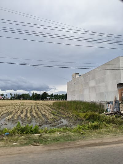 This image presents a wide view of a rural or semi-rural landscape in Grong-Grong, Indonesia, under an overcast sky. In the immediate foreground, a blurred dirt or unpaved road and green roadside vegetation suggest the photograph was taken from a moving vehicle.

The middle ground is dominated by a harvested rice paddy field, stretching horizontally across the frame. It features distinct rows of golden stubble interspersed with patches of muddy water, indicating recent harvesting and wet ground conditions. To the right, bordering the field, stands a large, multi-story, unpainted concrete building, likely commercial or industrial, with small windows or vents visible on its grey facade. A smaller structure is present on its rooftop. Next to this building, there's an area of tall green reeds and some accumulated debris, including corrugated metal and wooden planks.

In the background, a line of lush green trees, prominently featuring tropical palm trees, conceals most of a small cluster of houses with various roof colors. Beyond the trees, the faint silhouettes of distant mountains are visible beneath a heavy canopy of dark grey clouds that cover the entire sky. Numerous black power lines traverse the upper half of the image.

The scene conveys a muted atmosphere due to the heavily overcast weather, suggesting either impending rain or recently passed showers, as evidenced by the wet field. The time of day is daytime, but the dense cloud cover prevents direct sunlight. There are no people visible, and no discernible text appears in the image. The composition highlights a blend of traditional agriculture and new construction within a tropical setting.