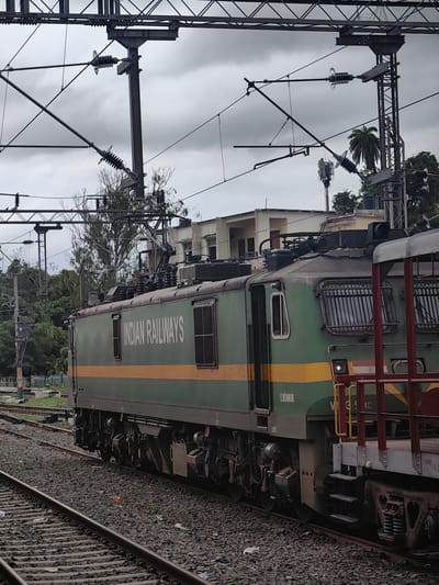 The image depicts a green and yellow Indian Railways locomotive at a train station in Khandwa, India. The locomotive is positioned on railway tracks, with multiple overhead electrical wires and supporting structures visible above. A portion of another railcar, painted red and white, is visible to the right of the locomotive. The background shows buildings, trees, and a cell tower, under a cloudy sky. The railway tracks are covered with ballast. The locomotive has the words "INDIAN RAILWAYS" printed on its side in white lettering. A portion of the locomotive's identification number, "WAG-9IC", is visible. The image is taken from a slightly elevated perspective, looking towards the side of the train.