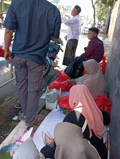 The image depicts an outdoor street scene in Pasuruan, Indonesia, under daylight conditions. The foreground features the back of a male subject wearing a dark blue short-sleeved shirt, gray cargo pants, and brown sandals, standing on a curb adjacent to a paved street. To the left, a portion of a wooden market stall is visible.
In the mid-ground, a blue motorcycle with a partially visible license plate (6163 JJM) is parked on the street. An older male, dressed in a white long-sleeved shirt, a patterned sarong, and a black peci, stands near the motorcycle, gesturing with his right hand. To his right, a younger individual with short dark hair, wearing a maroon long-sleeved shirt, is seated on a red plastic stool, smiling towards the left of the frame.
Further to the right, a white sheet is spread on the ground next to a concrete wall. Two female individuals are seated on the sheet, facing away from the camera. One wears a light brown or beige hijab, and the other wears a pink hijab over dark clothing, with a white strap visible. Around them are several red plastic bags containing various items, a white woven basket filled with green leafy vegetables, and bottled water. The ground shows some scattered debris. In the background, street signs are partially visible, and the scene extends down a street with other distant figures and structures.