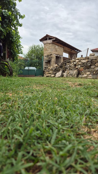 The image shows a low-angle view of a grassy yard with an overcast sky. In the background, there is a partial stone structure with a wooden roof, resembling ruins. To the left, a green metal gate is visible, with some greenery and what appears to be a clothesline with laundry hanging on it. A stone wall is also present to the right of the structure. The location is identified as Doirentsi, Bulgaria. The scene is devoid of any people or activity.