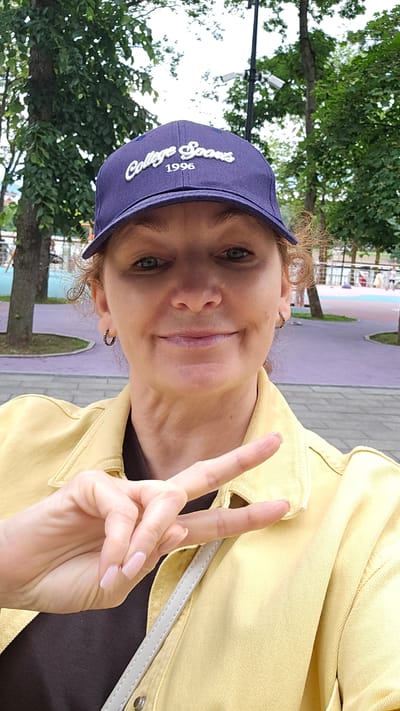 A woman wearing a purple baseball cap with "College Sooners 1996" embroidered on it, and a yellow jacket, is taking a selfie outdoors. She is making a peace sign with her right hand. She has curly hair, blue eyes, and is smiling. In the background, trees and a street with purple and pink paved paths are visible. The environment suggests an urban park or recreational area. There are security cameras mounted on a pole in the background.
