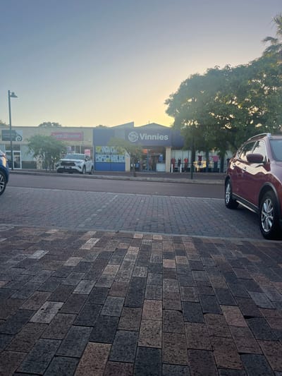 The image depicts a commercial street scene in Applecross, Western Australia, under the soft, warm light of either late afternoon or early morning. The immediate foreground shows a patterned paved surface composed of rectangular pavers in varying shades of grey, brown, and muted red. This paving transitions into a darker, asphalt road that runs horizontally across the middle ground.
On the road, a white Toyota Hilux Ute (pickup truck) with the license plate 1GSS460 is parked, facing left. Partially visible on the far left foreground is the front section of a blue vehicle. On the far right foreground, the side and front quarter of a red/burgundy SUV, likely a Nissan X-Trail, are visible.
Across the street, a row of single-story commercial buildings is visible. From left to right, these include a partially visible storefront with text appearing to read "CHARMED SALON", followed by a "Baskin Robbins" ice cream store featuring a pink logo. Adjacent to Baskin Robbins is a "Vinnies" (St Vincent de Paul Society) thrift store, identifiable by its prominent blue facade and white circular logo. The Vinnies store has large glass display windows showcasing various items, and external racks to the right of the entrance hold hanging clothes. A sign visible in one of the Vinnies windows reads "DONATE SHOP REPEAT". Several indistinct human figures are present inside and around the Vinnies entrance, suggesting customer or staff activity. Green trees are interspersed along the street, casting some shadows and partially obscuring the buildings and the bright, warm sky in the background. A single black street lamp pole is visible on the far left. The sky above is clear, transitioning from light blue to a yellowish glow near the horizon.