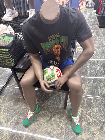 This image presents a retail display within a commercial establishment, likely situated in Sunrise, United States of America. The central focus is a headless, dark-skinned male mannequin seated on a black metal chair or bench. The mannequin is dressed in a dark gray short-sleeved t-shirt featuring a prominent "FIFA WORLD CUP" graphic in green and orange, an image of the FIFA World Cup trophy, and a partially legible list of host cities for the 2026 event including Vancouver, Monterrey, Los Angeles, Guadalajara, Dallas, Houston, Kansas City, Atlanta, Miami, Philadelphia, Foxborough, Seattle, San Francisco, and New York/New Jersey. The mannequin also wears royal blue athletic shorts and emerald green and crimson red athletic sneakers with white laces. Its hands cradle a white, green, and blue soccer ball, branded "VOYAGIA" and "TRIOMPHE," in its lap. A thin, translucent bracelet is visible on the mannequin's right wrist.

To the left, a black metal grid display unit holds several neatly stacked black t-shirts, identical to the one worn by the mannequin, with a white price tag attached to the topmost stack. In the background, on a similar black metal grid shelf, the lower legs and feet of two additional mannequins are visible, each displaying athletic shoes. One wears white sneakers with light blue Adidas stripes, and the other wears white sneakers with yellow laces. The flooring is composed of gray and white marbled tiles with visible grout lines, extending throughout the visible area of the store. Additional retail fixtures and blurred apparel are discernible in the deeper background.