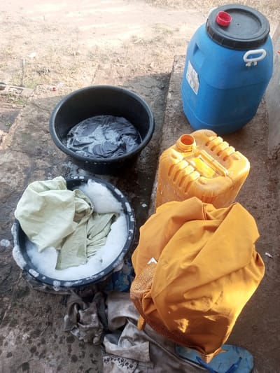 The image captures an outdoor laundry setup in Ebiya, Nigeria, under natural daylight. The scene is set on an uneven ground surface composed of concrete and dirt, with visible wet patches. Two black plastic washbasins are central to the foreground; the upper basin contains dark grey fabric submerged in white soapy water, while the lower basin holds light green checkered and solid light green fabrics also soaking in sudsy water. To the right, a blue plastic barrel with a black lid and a red cap is positioned upright next to an uncapped yellow plastic jerrycan. A large, crumpled piece of mustard yellow fabric rests on the ground near the lower right, partially covering a pair of light blue flip-flops. Several additional pieces of dull brown or grey fabric are scattered on the ground near the lower washbasin, one with a discernible dark pattern. No individuals are present in the frame.