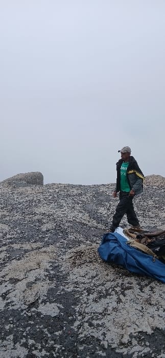 The image displays an individual on a rocky, high-altitude terrain at Gilman's Point, Tanzania, under a uniformly grey, overcast or foggy sky. An adult male, appearing in the mid-ground, wears a light-colored cap equipped with a headlamp, a dark jacket with yellow and grey details, a green t-shirt with a white 'adidas' logo, and dark trousers. He is observed in a walking or paused stance, facing left and slightly forward. The ground is characterized by irregular, textured grey and black rock, with larger rounded rock formations visible further back on the left. In the lower right foreground, a blue fabric item, possibly a rolled-up bag or tarp, is present along with other brown gear.