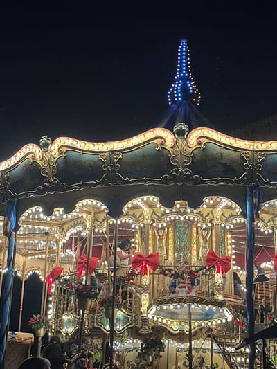 The image depicts a brightly illuminated, multi-tiered carousel at night in Santa Cruz Aquiahuac, Mexico. The carousel features elaborate golden scrollwork, light-colored panels, and numerous warm white incandescent bulbs outlining its structure and decorative elements. The upper band of the carousel is dark blue/green with painted scenic vignettes, possibly depicting bridges and cityscapes. The carousel's conical roof is dark blue, topped with a spire-like structure illuminated by blue lights, resembling the Eiffel Tower. Large red bows with green and red floral garlands adorn various sections of the carousel.
Several individuals are present on the carousel. On an upper tier, a woman with dark hair, wearing a light-colored top and sunglasses, is visible, appearing to stand or sit on a stationary platform, looking towards her left. To her left, two raised arms are partially visible near a red bow. In the center of a lower tier, a doll or figurine dressed in blue and white, wearing a hat and holding a package, is displayed. Portions of other people, including what appears to be a child on a white carousel animal, are visible in the lower section of the frame. The immediate environment is illuminated by the carousel's lights, while the background beyond the carousel is dark, indicating nighttime.