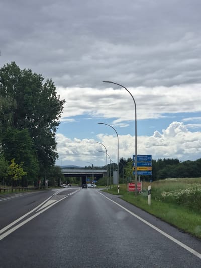 The image captures a view from a vehicle traveling on a wet asphalt road, likely on a cloudy day, with large trees lining the left side of the road. On the right side, there are fields of green grass and wildflowers. Overhead, a series of modern streetlights follow the curve of the road. In the distance, a concrete overpass supports traffic. A collection of road signs are visible on the right, indicating directions to Saarbrucken, Saarlouis, Schengen via A13, Remich via A13, Mondorf via N16, and Luxembourg/Aspelt. One sign also indicates a detour ("Deviation") for Mondorf via A13. Several vehicles are present on the road, including a black car, a white van, and other cars in the distance. The overall environment suggests a semi-rural or highway setting.