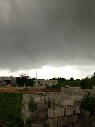 This image captures an outdoor scene in Ajerogun, Nigeria, under heavily overcast, dark grey storm clouds that dominate the sky. In the foreground, multiple stacks of weathered, hollow-core concrete blocks are visible, partially overgrown with green vegetation. The immediate ground consists of reddish-brown soil mixed with patches of green grass.

In the midground, to the left, a partially completed, single-story building with unrendered grey walls and two rectangular windows is present. An open doorway leads to a small porch, where a blue bucket and a white container are visible. Centrally, a tall, slender, light-colored pole stands vertically. Further right, an area of low concrete block foundations or partial walls is visible, upon one of which rests a metallic pot, possibly silver, with a red lid or handle.

The background consists of dense green foliage and trees, some appearing to be swaying or leaning due to wind. No human figures are present in the scene. The overall environment suggests an undeveloped or active construction site experiencing blustery, pre-storm conditions.