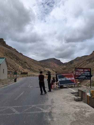 The image captures a road checkpoint in a mountainous, sparsely vegetated region under a cloudy sky. In the foreground, a paved road stretches into the distance, flanked by dry, rocky hillsides. Three individuals are present near the road. Two men stand on the right side of the road, facing left, while a third person sits on a chair on the left side. A prominent sign on the right reads "THANK YOU HAVE A SAFE JOURNEY DISTRICT POLICE LEH LADAKH FOR EMERGENCY DIAL 112". Another sign, partially visible, displays "DISTRICT POLICE.LEH". A small, light-colored building with a green roof is situated on the left side of the image. The environment suggests a remote location, possibly a border area or a high-altitude pass. The presence of the police signs and personnel indicates an official checkpoint.