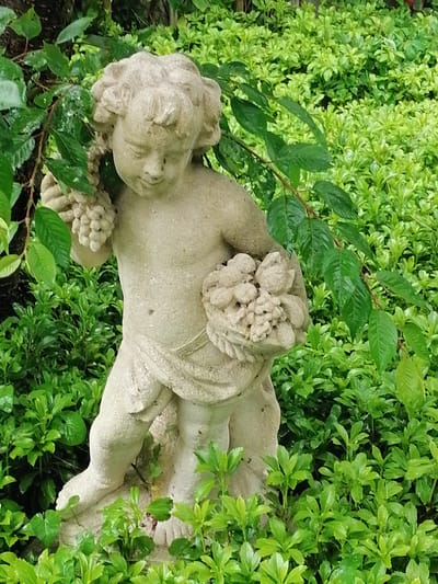 The image depicts a stone statue of a cherub in a garden setting. The statue is positioned in the center of the frame, surrounded by lush green foliage. The cherub is rendered in a light-colored stone, with curly hair and a serene expression. It holds a cluster of grapes in its left arm and a basket overflowing with what appears to be fruit or flowers in its right hand. The garden is densely planted with small, leafy shrubs, creating a vibrant green backdrop. Overhanging branches with dark green leaves partially obscure the statue, adding depth and a sense of natural integration. The lighting suggests it is daytime, with diffused light indicating possible cloud cover or shade. The overall scene conveys a tranquil and idyllic garden atmosphere. The location is identified as Grünwald, Germany, though no specific landmarks or identifying features of the city are visible within the frame itself.