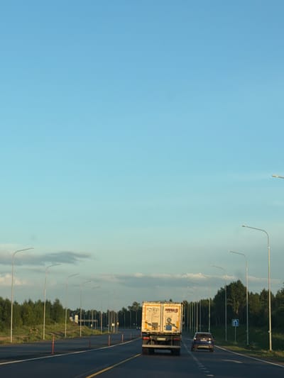 The image shows a view from a vehicle traveling on a highway, possibly in or near Kaipy, Russia, during daylight. The sky is a clear, pale blue with some wispy clouds. On the right side of the highway, a line of tall streetlights stretches into the distance, flanking a dense line of green trees. The highway itself is asphalt with yellow and white lane markings.

In the foreground, a white semi-trailer truck with a box trailer is traveling in the right lane. The trailer has some markings on it, including what appears to be a stylized face and the text "MEGA 3 LIGHTS". A dark blue car is in the left lane, following the truck. Orange traffic cones are visible along the side of the road. Further down the highway on the right, a blue and white road sign with an arrow indicating a turn is present. The overall impression is of a modern highway with well-maintained infrastructure, set against a natural backdrop of trees and a clear sky.