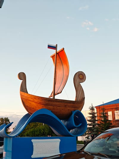 The image depicts a monument resembling a historical wooden boat, possibly a longship, situated on a blue wave-like pedestal in Nikolo-Beryozovka, Russia. The boat features a distinct, upward-curving bow and stern, with the stern adorned by a painted yellow sun symbol. A single red-orange sail is hoisted on a mast, which is topped with a Russian flag (white, blue, red horizontal stripes). The boat rests on a layered, sculptural blue base designed to mimic ocean waves. The base includes a white scroll-shaped plaque containing dark text, likely an inscription, facing the viewer.
The scene is illuminated by the soft, warm light of late afternoon or early evening, casting a glow on the monument and the surrounding environment. The sky above is a light, clear blue with a few scattered, wispy white clouds. In the background to the right, a two-story red-brick building with a dark blue corrugated metal roof is visible, featuring several white-framed windows. Two coniferous trees stand in front of this building. To the left, beyond the monument, lush green foliage is discernible.
In the mid-ground, a person wearing a dark shirt is seen standing next to a dark SUV, partially obscured by the monument and the foreground. Another person's head is faintly visible through an opening in the blue wave structure on the left side of the monument. The foreground is occupied by the dark hood and windshield of a car, reflecting portions of the surrounding scene and other vehicles.