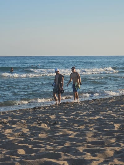 The image, taken at Castelldefels, Spain, shows a beach scene with two people visible. The perspective is from a high angle, looking down at the shoreline. The right side of the frame is dominated by textured sand, likely from the dunes of Castelldefels beach, with distinct shadows indicating a low sun angle, suggesting either early morning or late afternoon.

To the left, the shallow, blue water of the Mediterranean Sea is visible, with small waves breaking near the shore. The sky is a pale blue, transitioning to a soft white near the horizon, indicating clear weather.

In the water, near the left edge of the frame and partially obscured by waves, is a person. Closer to the shore, standing at the water's edge with their feet in the shallow water, is another person. This individual is wearing swim trunks and appears to be holding a light-colored towel or piece of fabric. Their posture is relaxed. Further out in the water, several small, dark objects can be seen, possibly buoys or other beachgoers.

The overall mood of the image is peaceful and leisurely, capturing a moment of relaxation by the sea. There is no visible text in the image.