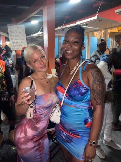 Two smiling women pose at an outdoor bar. The woman at the top has an exposed midriff, blue dress, shoulder tattoo, and gold chain. The woman at the bottom has a blonde bun, and a long palm tree-print dress and holds a drink with a black straw. They are standing near the "Tipsy Bar" menu. People are scattered in the background.