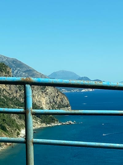 The image shows a view of the Adriatic Sea and a coastline from a balcony or bridge in Seoce, Montenegro. In the foreground, a weathered blue metal railing with visible rust runs horizontally across the frame, with vertical posts supporting it. Beyond the railing, the deep blue sea stretches to the horizon under a clear, light blue sky. The coastline features rugged, steep cliffs covered in green vegetation. Several small white boats are visible on the water, some leaving white wakes. In the distance, hazy mountains rise, their peaks softened by the atmospheric perspective. The overall scene depicts a sunny day with a scenic coastal landscape.