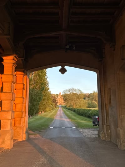 The image captures a view looking out from a dark, ornate stone archway onto a long, tree-lined driveway that recedes into the distance, leading to a grand building. In the immediate foreground, the interior of the archway features dark wooden structures, possibly ceiling beams or window frames, and carved stone elements, including a distinctive finial. Below the opening, a low stone wall or parapet glows with warm, orange light from the low sun.
Beyond the archway, a paved driveway stretches forward, bordered on both sides by manicured green lawns and tall, dense trees. A dark-colored vehicle, possibly an SUV, is partially visible on the right side of the driveway. The focal point in the far distance is a large, stately building with intricate architecture, likely a historic country house or castle, constructed from light-colored stone. The sky above is clear and light blue.
The scene is bathed in the warm glow of the golden hour, indicative of either late afternoon or early morning, with the low sun casting long shadows across the driveway and intensely illuminating the foreground stone with a rich, amber hue. There are no people visible, and no discernible text. The setting evokes a sense of tranquil grandeur, consistent with a large estate in the South Kesteven, United Kingdom area.