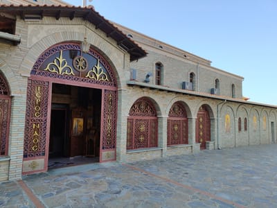 The image shows a side view of the exterior of the Church of Agios Nikolaos in Litochoro, Greece. The church has a facade constructed with light-colored bricks and stone. The main entrance is a large arched doorway with decorative metal gates. Above the entrance, there is an inscription in Greek letters. To the right of the entrance, a series of arched windows are covered with similar decorative metal grilles. The building extends to the right, with a wall featuring several arched niches containing religious icons. The ground in front of the church is paved with irregular flagstones. The sky is clear and blue, and the lighting suggests it is daytime. There are no people visible in the image.