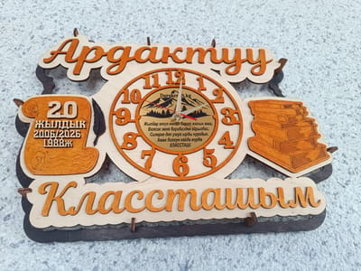 This is a medium shot, eye-level, close-up image of a wooden plaque designed to look like a clock and a stack of books, with decorative text. The plaque is mounted on a textured, light-colored surface that appears to be a wall with a subtle glitter effect.
The central element is a round clock face with orange numbers and stylized wooden hands. The background of the clock face features a printed image of mountains and a dark sky. Above the clock, large, orange, stylized letters spell out "Ардактуу," which translates to "Dear" in Kazakh. To the left of the clock, there is a wooden cutout shaped like a scroll, containing the number "20" in large orange font, followed by smaller text reading "ЖЫЛДЫК 2006/2026 1988ж." This likely refers to a period of 20 years, perhaps a class reunion, with specific years noted.
To the right of the clock is a wooden carving of a stack of books, also in orange. Below the clock and the surrounding elements, a large, wavy, orange banner contains the word "Классташым," which translates to "My Classmate" in Kazakh.
The overall aesthetic is commemorative and nostalgic, likely a gift or memento for classmates. The image is well-lit, highlighting the details and the natural wood textures. There are no people visible in the image, focusing entirely on the handcrafted object. The background suggests an indoor setting or a display area, as the textured wall is a consistent backdrop.