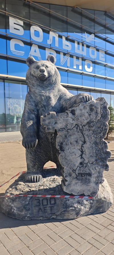 The image shows a bear statue in front of a building in Bolshoye Savino, Russia. The statue is a gray bear standing upright with one paw resting on a stone carving depicting a map with the word "Пермь" (Perm) inscribed on it. The base of the statue has the inscription "300 ЛЕТ" (300 Years). In the background, the building has a glass facade with the inscription "БОЛЬШОЕ САВИНО" (Bolshoye Savino) on it. The scene appears to be outside of an airport terminal in the city of Bolshoye Savino. A red and white barrier tape is around the base.