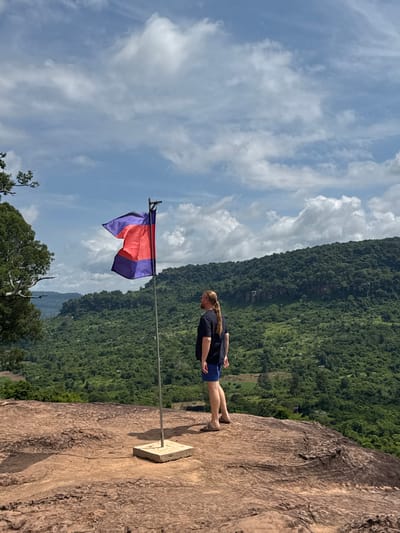 In Banteay Srei, Cambodia, a person stands on a rocky outcrop overlooking a lush green valley. The person, viewed from behind, has long blonde hair tied in a ponytail and is wearing a dark t-shirt and blue shorts. A flag, horizontally striped with purple, red, and purple, flutters on a pole in front of them. The background features a dense, tree-covered mountainside under a sky with scattered white clouds.
