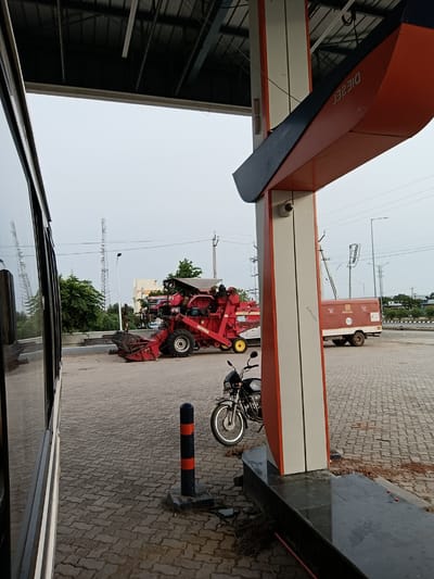 In Sugalimetta, India, a red combine harvester is parked on a paved surface under a canopy at a fuel station. To the right of the harvester, a red and white utility vehicle is parked. A black motorcycle is parked in front of a fuel pump structure, which has an orange and black design and a security camera mounted on its side. The ground is paved with interlocking bricks. A large vehicle, possibly a bus, is partially visible on the left side of the frame, reflecting the surroundings. In the background, communication towers, trees, and buildings are visible. The sky is overcast.