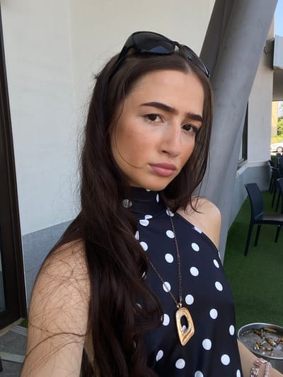 An outdoor selfie is taken from a medium close-up perspective, featuring a young woman with long, dark hair, and fair skin. She is wearing a black, halter-neck dress with a white polka dot pattern, a gold pendant necklace, and sunglasses perched on her head. Her gaze is directed towards the camera, with parted lips. In the background, to the right, an ashtray overflowing with cigarette butts is visible, alongside a metal table and several black chairs arranged on a green, artificial turf surface. The scene appears to be an outdoor seating area of a cafe or restaurant in Somma Lombardo, Italy.