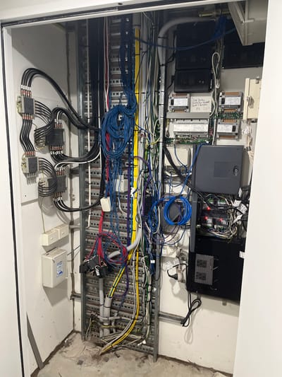 This image captures the interior of an open electrical or data control cabinet, showcasing a complex array of wiring and electronic components. The cabinet appears to be part of an industrial control system or a building management system, located in a utility or plant room in Kelvin Grove, Australia.
Numerous electronic modules, resembling PLCs (Programmable Logic Controllers) or I/O modules, are mounted on DIN rails across the upper and middle sections of the cabinet. These include several grey and black units, alongside what appear to be power supplies and network devices. A central grey module has partially legible text, including "Sac Jet 6x 6x...".
A dense network of cables, in various colors such as blue, white, black, yellow, red, green, and purple, runs throughout the cabinet. Many cables are routed within metal cable trays or ladders, while others, especially bundles of blue data cables, form loose coils. Small white labels provide identification for numerous cables and modules, featuring codes like "CV-1", "CV-2", "CV-3", "FCU-1" through "FCU-10", and "FX-1" through "FX-4". These likely refer to Control Valves, Fan Coil Units, and field expanders, strongly indicating a HVAC or building automation application.
The lower part of the cabinet displays several identical black rectangular components, possibly current transformers or terminal blocks, meticulously wired with smaller black cables. Adjacent to these are a small white rectangular box and a larger white box with a clear lid, marked with "IP65", signifying its ingress protection rating. The background on the right side reveals an exposed, unfinished wall surface, likely concrete, consistent with a technical service area. No human subjects or activity are present; the image documents the static state of the electrical installation under artificial lighting.