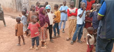 The image depicts approximately 20-25 individuals, predominantly children and young adolescents, gathered outdoors on a reddish-brown dirt surface in Rigasa, Nigeria. In the foreground, a young child wearing a red t-shirt and dark pants stands to the left of center, looking right. Another young child in an orange patterned shirt stands with their back to the viewer, facing left. Several other children are positioned behind them, many wearing sandals or flip-flops, some barefoot.
To the left, a young girl in a dark purple hijab and a floral dress directly observes the viewer. Adjacent to her, a boy in a light yellow long-sleeved shirt and blue jeans looks toward the right. Other children wear various attire including light blue caftans, red long-sleeved shirts, and light-colored t-shirts. Many children appear to be observing something or someone to the right of the frame. One boy in a light blue collared shirt and blue jeans has his right hand near his mouth.
On the far right, an adult male, partially visible from the chest down, wears a blue polo shirt with a yellow collar trim and a dark vest over blue jeans. He holds a dark camera or similar device, facing away from the viewer, suggesting he is engaged in photography or videography.
The background consists of a weathered, light brown wall, possibly mud or brick, with a dark wooden door visible behind some of the children on the left. The ground is consistently unpaved dirt. The scene is illuminated by diffused daylight.