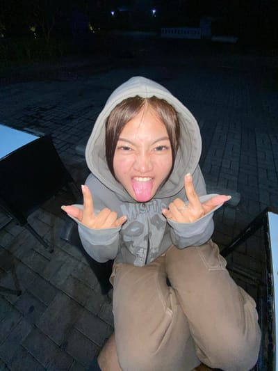 A young woman, with long dark hair, wearing a grey hooded sweatshirt and tan cargo pants, sticks her tongue out at the camera. She is making "rock on" hand gestures with both hands. She is seated outdoors at night on a patio with brick paving. In the background, there are dimly lit trees and buildings. A black chair is visible to her left.