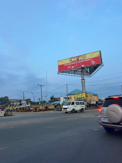 The image shows a street scene in Ojoto, Nigeria, during what appears to be the early evening or late afternoon. A large billboard with a red and yellow design is prominently displayed above a street intersection. The billboard features text that reads "MANY DREAM ABOUT IT, FEW WORK TOWARDS IT. BE YOUR BEST." and includes images of athletes. Below the billboard, a yellow building with a blue roof houses a business with signage that includes "AUSTONE TIRES."
Several vehicles are present, including a white van driving on the road, a silver SUV with its brake lights illuminated, and numerous auto-rickshaws (popularly known as "keke napep") parked or driving along the side of the road. A bus is partially visible on the left side of the image. A group of people are gathered near the auto-rickshaws, and some individuals are on motorcycles. In the background, buildings, streetlights, and utility poles are visible, as well as a tall communication tower. The sky is overcast with a light blue hue. Another billboard on the left side of the street has text that reads "GENDER PROGRAMME... TO SHUT UP". The overall impression is that of a busy urban thoroughfare.