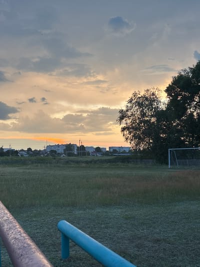 The image depicts an outdoor scene, likely at dusk, with a sky filled with clouds tinged with orange and yellow hues from the setting sun. In the distance, a low-rise urban or semi-urban landscape is visible, featuring several buildings, some appearing to be apartment blocks, and smaller houses. A fence runs across the middle ground. To the right, a football goal is partially visible. The foreground is dominated by a grassy field, with what appear to be parallel bars or railings in the lower portion of the frame, one metallic and rust-colored, the other painted blue. The overall impression is one of a park or recreational area at the edge of a residential zone, with the natural light of sunset casting a warm glow. The location is identified as Sarootski Selski Savet, Belarus. There are no people or animals clearly visible in the image.