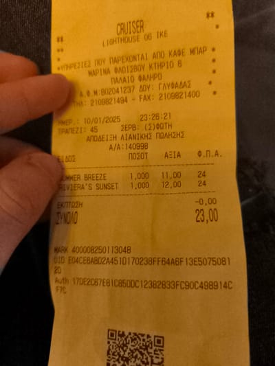 A person's fingers hold a yellow paper receipt from a business called "CRUISER LIGHTHOUSE 06 IKE." The receipt is presented against a dark, textured background, likely clothing.

The receipt documents a retail transaction that occurred on 10/01/2025 at 23:26:21. The location is "MARINA FLOISVOU KTHRIO 6, PALAIO FALIRO," which is in Palaio Faliro, Greece. The tax office (DOU) is listed as Glyfada, and the VAT ID (A.F.M.) is 802041237. Contact numbers are also provided (Tel: 2109821494, Fax: 2109821400). The server for table 45 is identified as Foti.

Two items are listed: "SUMMER BREEZE" for 11.00 and "RIVIERA'S SUNSET" for 12.00, each with a quantity of 1.000 and a VAT rate of 24%. There was no discount applied. The total amount due is 23.00.

Additional details on the receipt include a MARK number (400008250113048), a UID (E04CE6AB02A451D170238FF64A6F13E5075081), and an authorization code (Auth 17DE2C67E8IC85DDC12382833FC90C498914CF7C). A QR code is visible at the bottom of the receipt. The Greek text at the top, "ΥΠΗΡΕΣΙΕΣ ΠΟΥ ΠΑΡΕΧΟΝΤΑΙ ΑΠΟ ΚΑΦΕ ΜΠΑΡ," indicates "Services provided by a cafe bar."