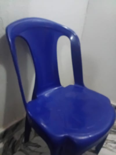 The image shows a bright blue plastic chair with a rounded seat and back, and armrests that curve downwards to form part of the legs. The chair is positioned at an angle, with its seat facing towards the viewer and slightly to the right. The background is a plain, light-colored wall, with a patterned fabric visible to the right of the chair, partially obscuring it. The lighting appears to be indoor, with a soft, even illumination. There is no activity or event taking place, and no people are visible. The image is a close-up shot of the chair, focusing on its form and color. The location context of Pantinapu, Nigeria, does not provide any further visual cues within the image itself. There is no visible text in the image.