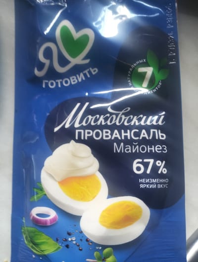 The image displays the packaging of a mayonnaise product. The dominant color of the packaging is blue. In the upper left corner, a white stylized heart symbol with a green fill is visible, accompanied by the Cyrillic text "Я ГОТОВИТЬ" which translates to "I am ready to cook." To the right of this, a dark circular emblem contains the number "7" within a green leaf-like design, surrounded by Cyrillic text indicating "NATURAL INGREDIENTS." The central and largest text on the packaging reads "Московский ПРОВАНСАЛЬ Майонез," meaning "Moscow Provence Mayonnaise." Below this, the percentage "67%" is prominently displayed, followed by the Cyrillic text "НЕИЗМЕННО ЯРКИЙ ВКУС" which translates to "Invariably Bright Taste." The bottom portion of the packaging features illustrations of halved hard-boiled eggs with mayonnaise, a slice of red onion, and green leaves, suggesting the ingredients or presentation of the product. The scene does not provide any information or context related to the city of Cajba, Moldova.