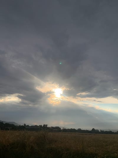 A wide, vertical shot captures a dramatic sky over a rural landscape at dusk. The sky is dominated by heavy, dark gray clouds that obscure much of the sun, but shafts of light break through, illuminating a patch of sky in the upper center with a bright, yellowish glow. These light beams fan out and create a spotlight effect on the clouds and a hazy atmosphere. In the distance, silhouetted against the lighter sky, are rolling hills or mountains, and a low-lying town or village with numerous buildings and trees. In the foreground, a field of dry, golden-brown grass or crops stretches across the bottom of the frame, its texture detailed but muted by the low light. The overall impression is one of natural drama, possibly a storm passing or approaching. The location is identified as Somoniyon, Tajikistan.