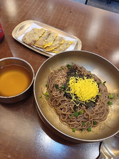 The image captures a Korean meal served on a dark-brown, textured table inside a restaurant in Mapo-gu, South Korea. Dominating the lower-right is a brass-colored metal bowl filled with buckwheat noodles (likely *makguksu* or *memil-guksu*), garnished with shredded yellow egg (jidan), chopped green onions, dark seaweed flakes, and sesame seeds. To the left, a smaller, matching metal bowl contains a light amber-colored broth. In the upper-left, a rectangular gold-toned tray holds five pieces of pan-fried meat jeon (*yukjeon*), each featuring a yellowish egg coating and arranged on white parchment paper. A traditional metallic Korean spoon is partially visible at the bottom-right of the frame. A red container or bottle is partially obscured on the far-left edge of the table.